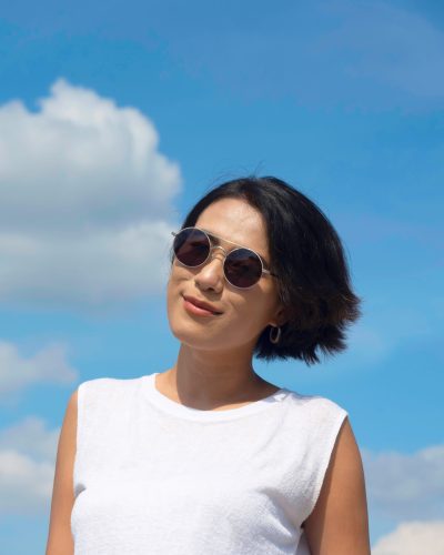 Happy woman in summertime. Smiling beautiful Asian woman short hair wearing sunglasses and casual white sleeveless shirt looking at camera on blue sky and cloud background on sunny day in summer.