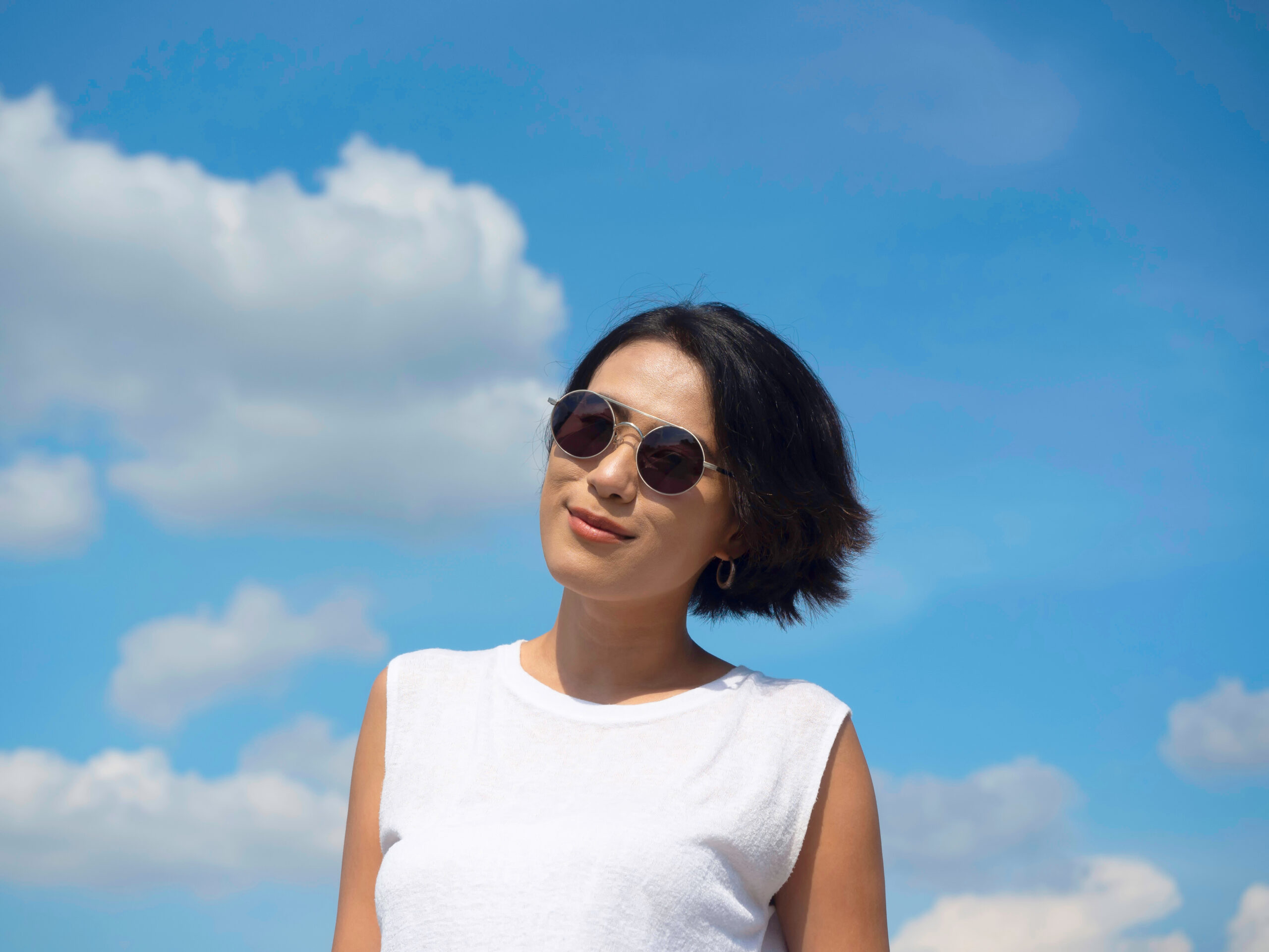 Happy woman in summertime. Smiling beautiful Asian woman short hair wearing sunglasses and casual white sleeveless shirt looking at camera on blue sky and cloud background on sunny day in summer.
